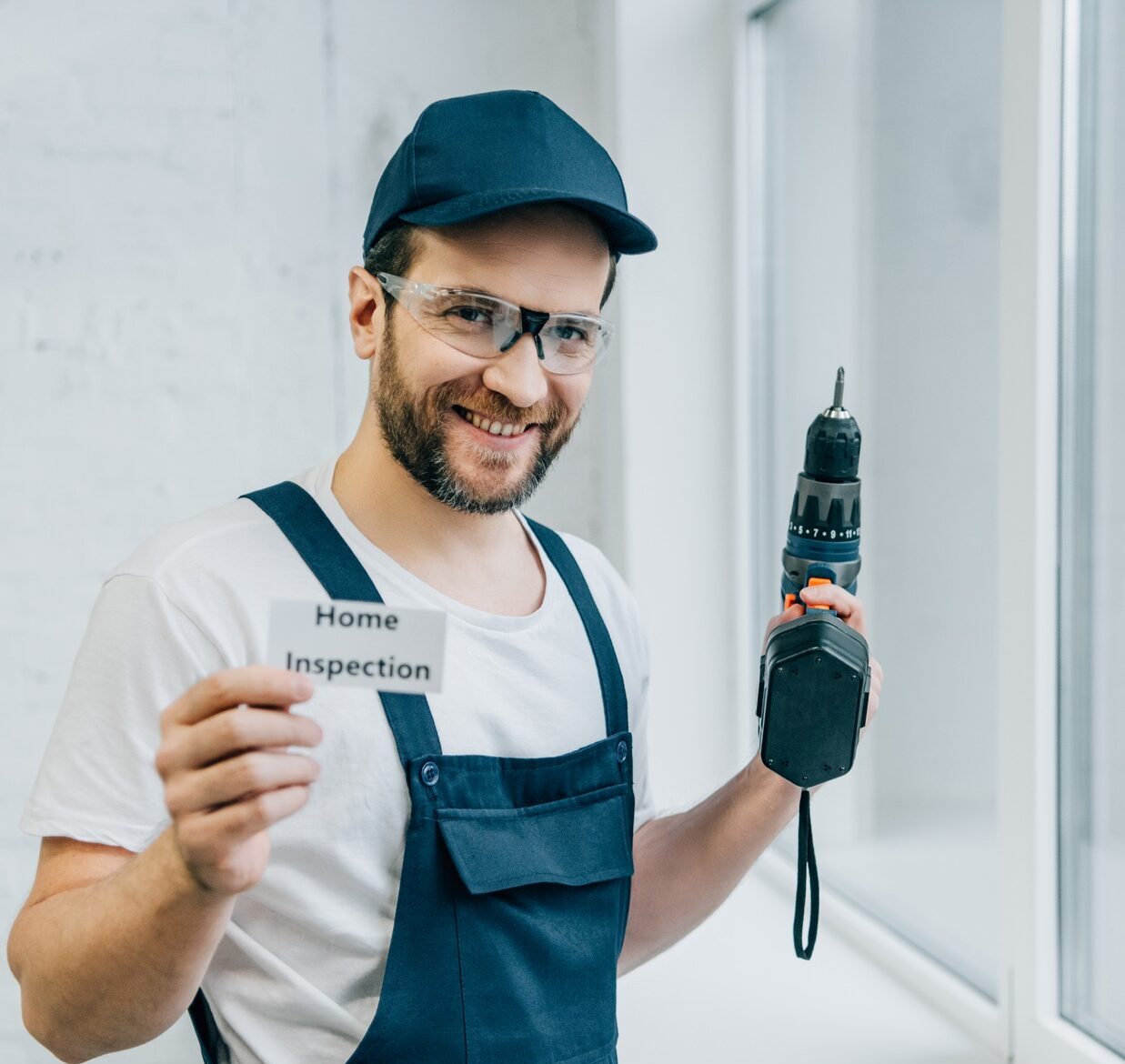 happy adult male craftsman holding electric drill and showing card with lettering home inspection